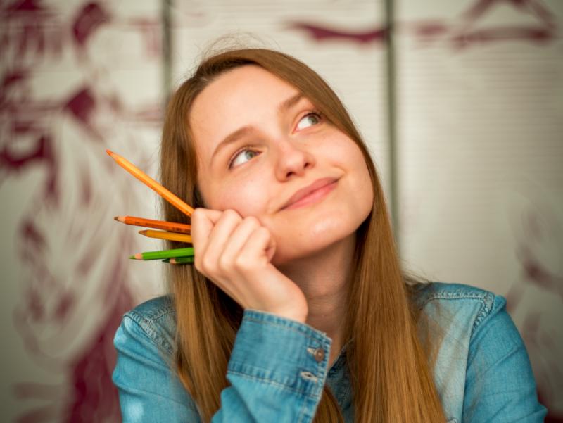 Eine Frau stützt ihr Kinn mit der Hand ab, in der sie Bleistifte in verschiedenen Farben hält. Sie blickt fröhlich, nachdenklich.