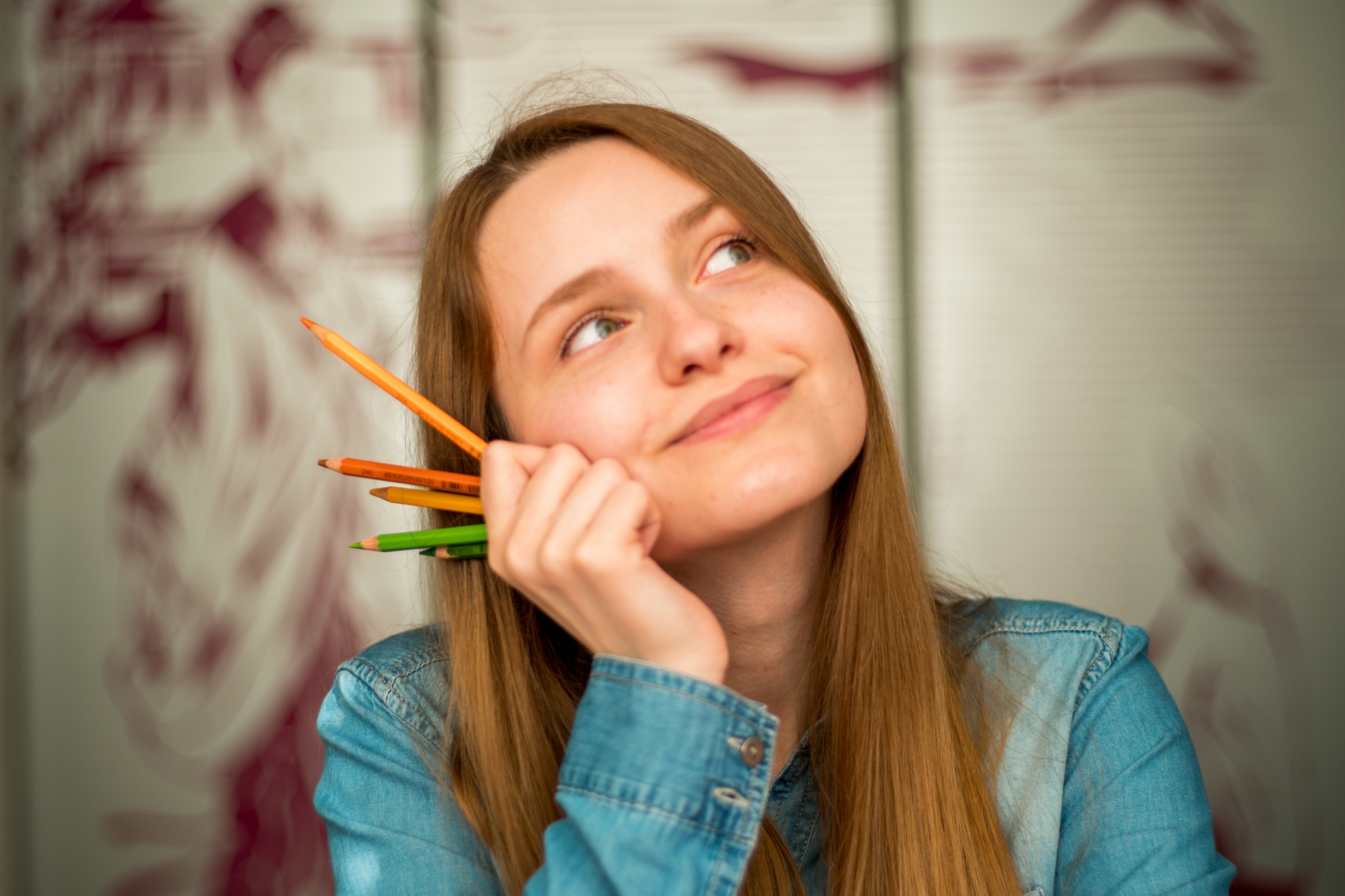 Eine Frau stützt ihr Kinn mit der Hand ab, in der sie Bleistifte in verschiedenen Farben hält. Sie blickt fröhlich, nachdenklich.