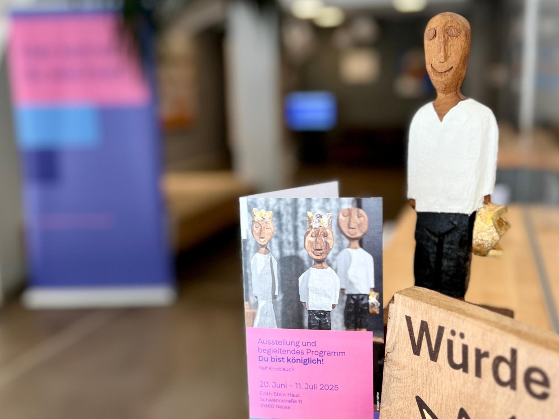Holzskulptur eines Königs mit Krone in der Hand, Broschüre zur Auststellung und ein Holzschild mit der Aufschrift 'Würde unantastbar'