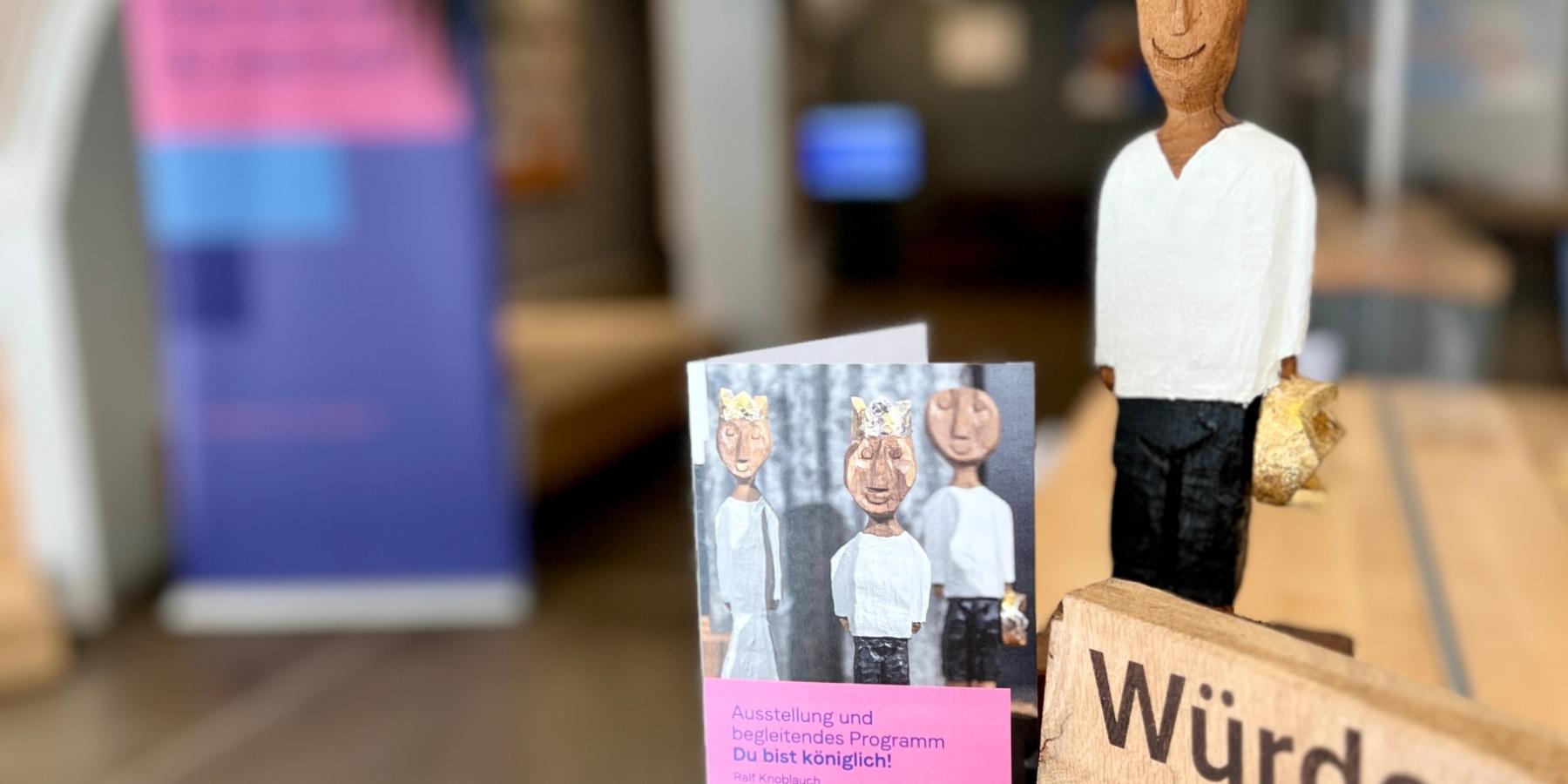 Holzskulptur eines Königs mit Krone in der Hand, Broschüre zur Auststellung und ein Holzschild mit der Aufschrift 'Würde unantastbar'