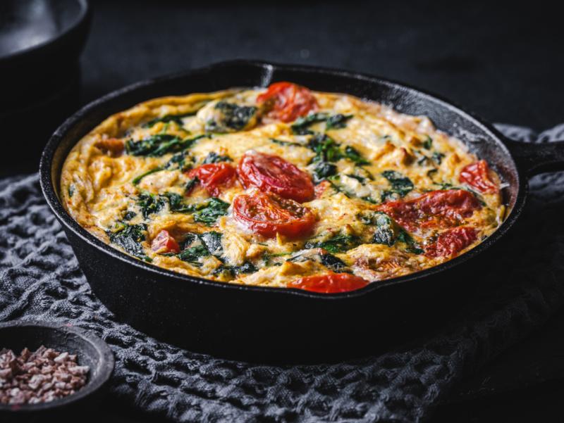 Überbackenes Gericht mit Spinat und Tomaten in einer gusseisernen Pfanne vor schwarzem Hintergrund.