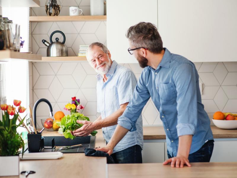 Zwei Männer unterhalten sich einander zugewandt in einer Küche. Ein von ihnen wäscht gerade Salat an einer Spüle.