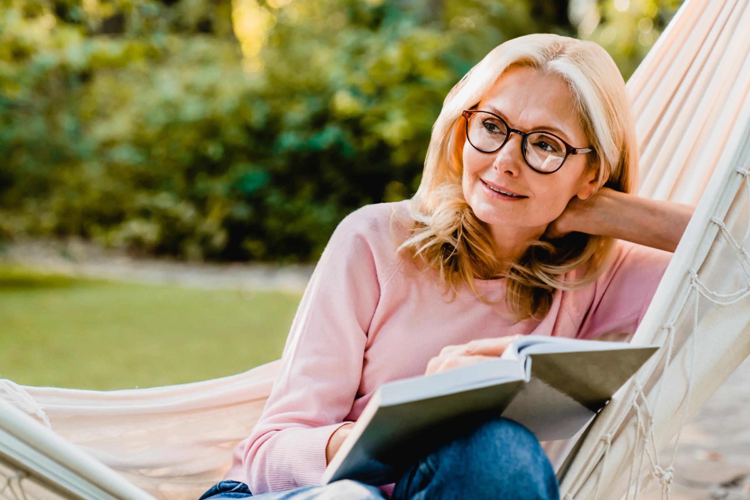Eine Frau sitzt in einer Hängematte, hat ein aufgeschlagenes Buch auf ihren Beinen liegen und schaut in die Ferne.