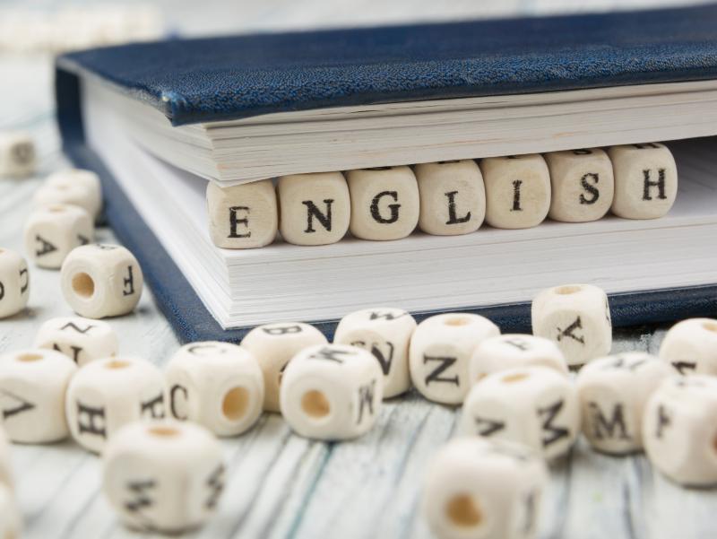 Das Wort english liegt aus Blockholzbuchstaben gebildet zwischen den Seiten eines geschlossenen Buches. Über den Holztisch, auf dem das Buch liegt, verteilt liegen weitere Blockholzbuchstaben.