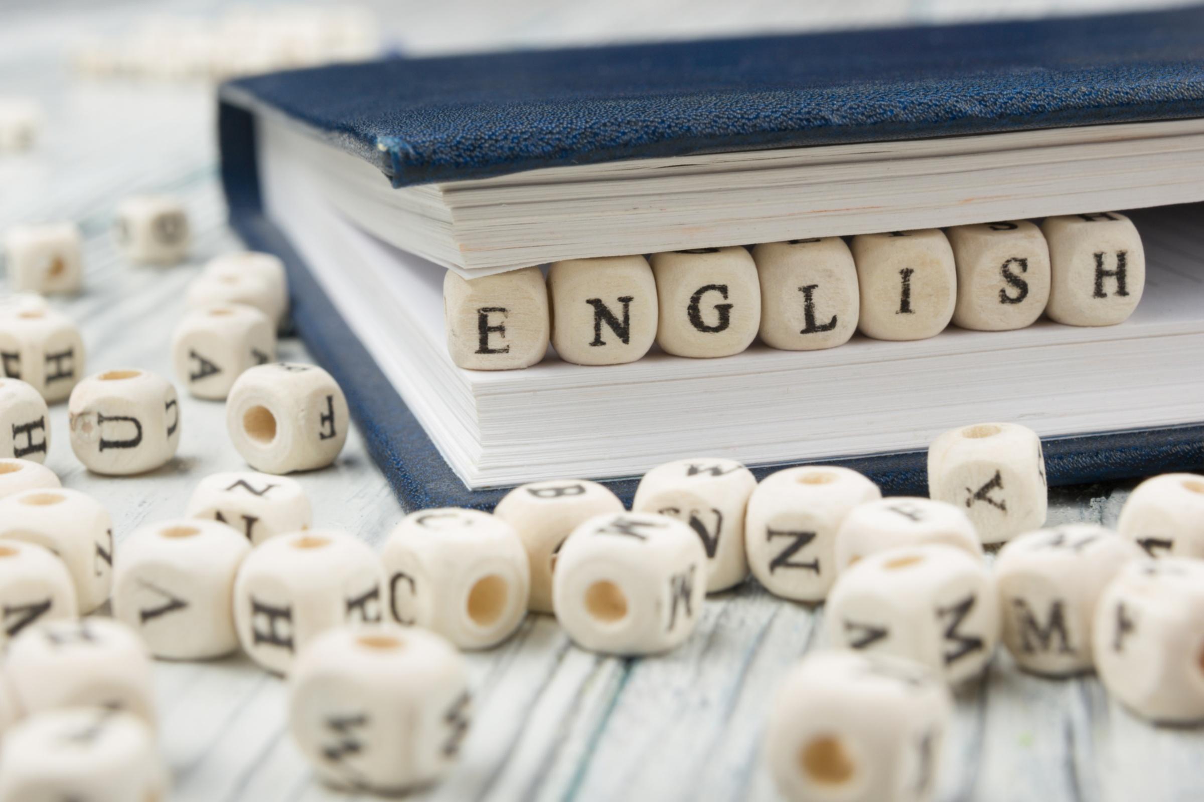 Das Wort english liegt aus Blockholzbuchstaben gebildet zwischen den Seiten eines geschlossenen Buches. Über den Holztisch, auf dem das Buch liegt, verteilt liegen weitere Blockholzbuchstaben.