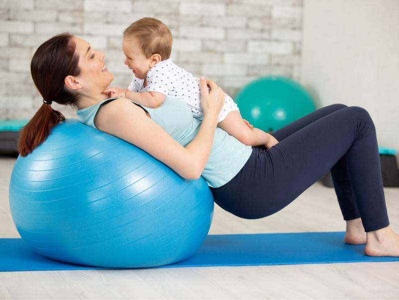 Die Mutter liegt für eine sportliche Übung mit dem Rücken auf einem Gymnastikball. Ihr Baby liegt auf ihr. Beide schauen sich fröhlich an.