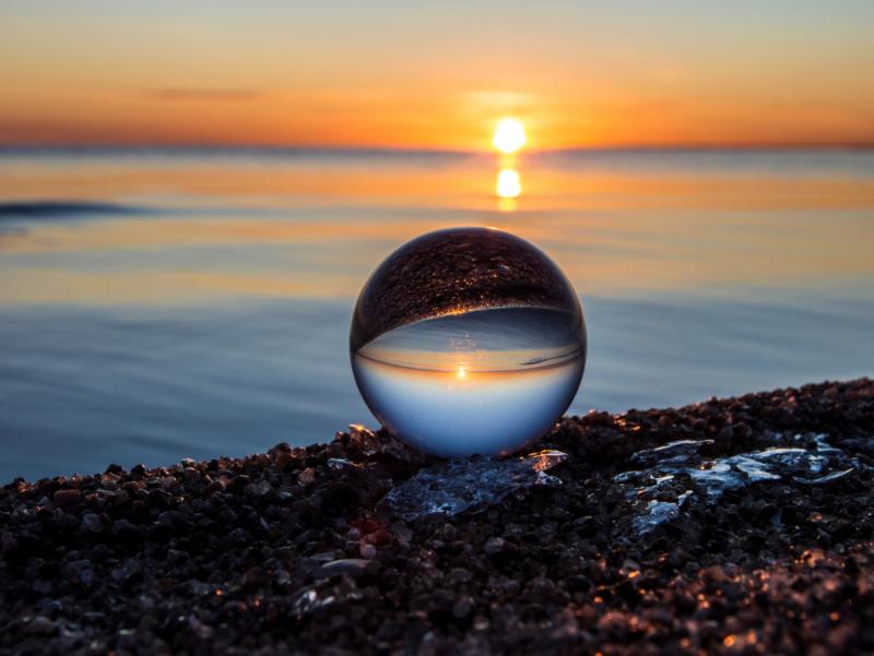 Eine Galskugel liegt am Ufer eines Kieselstrandes. Durch sie hindurch ist der Sonnenuntergang zu sehen.