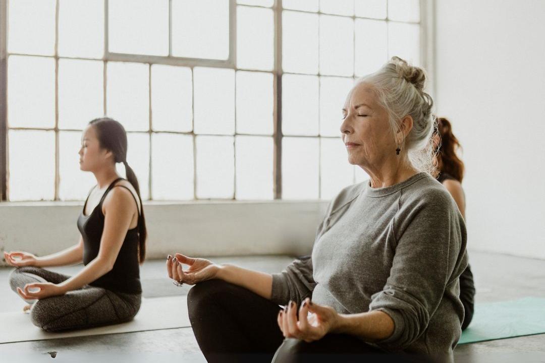 Drei Frauen sitzen in einer Yoga-Position in einem hellen Sportraum.