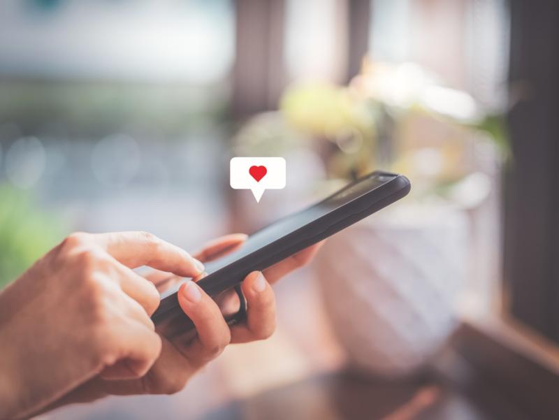 Hand einer Frau, die ein Smartphone bedient, vor dem Hintergrund eines Cafés. Über dem Handy erscheint ein Herz-Symbol.