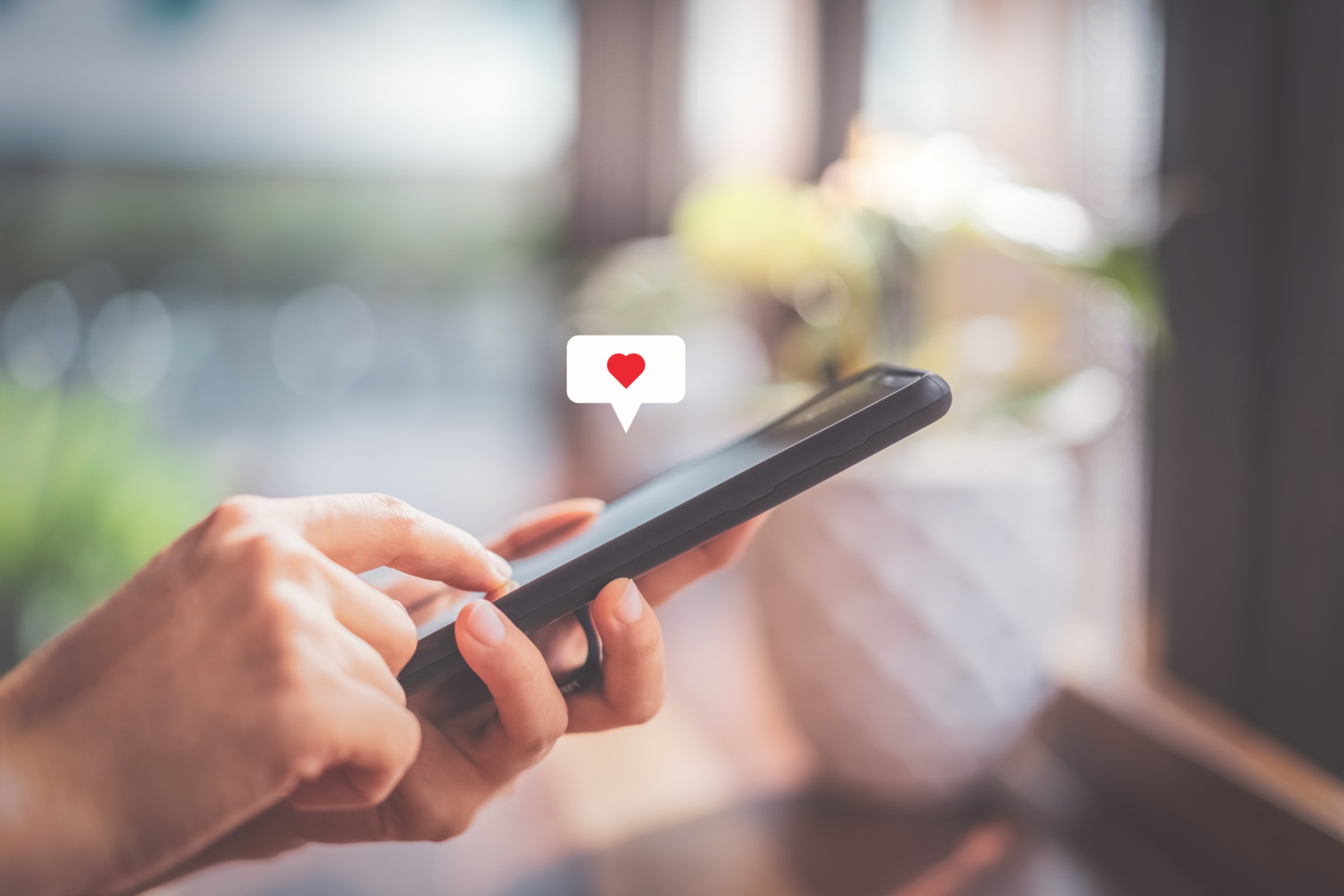 Hand einer Frau, die ein Smartphone bedient, vor dem Hintergrund eines Cafés. Über dem Handy erscheint ein Herz-Symbol.