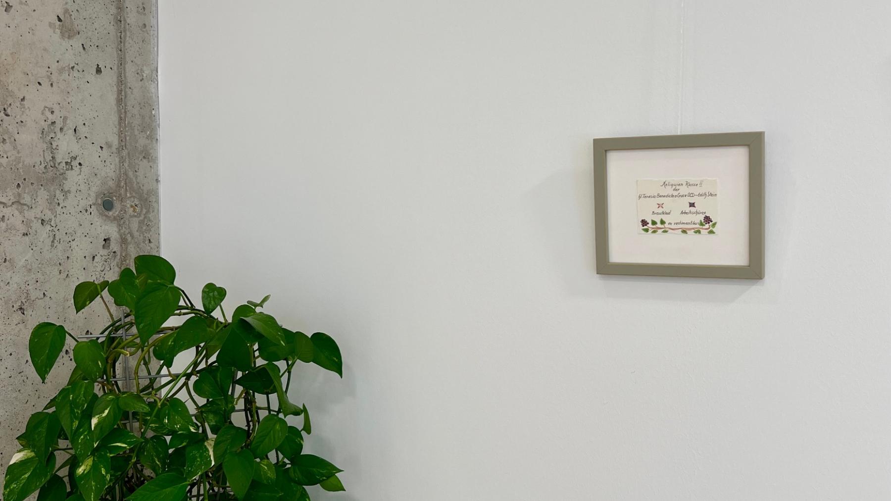 In einem Rahmen hängt an der Wand im Raum des Stille ein Karte auf denen zwei kleine Stoffstücke aufgenäht sind. Reliquien von Edith Stein: Ein Teil ihres Brautkleides, mit dem sie beim Eintritt ins Kloster gekleidet war und ein Teil ihrer Arbeitsschürze.